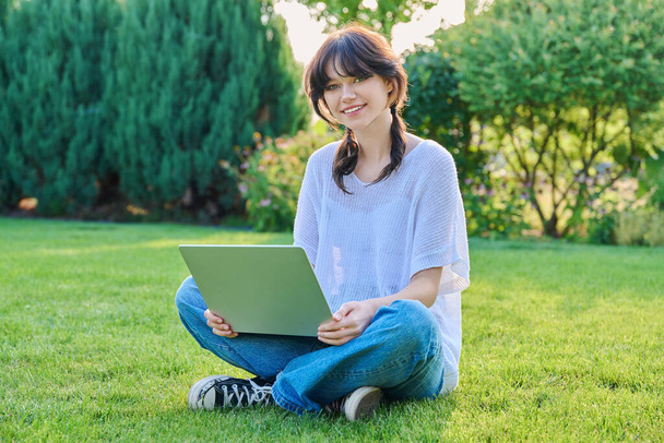 Teenage girl sitting on grass using laptop. Female teenager student 18, 19 years old sitting on lawn, looking at camera, technologies for studying leisure communication shopping in backyard in garden - Photo, image