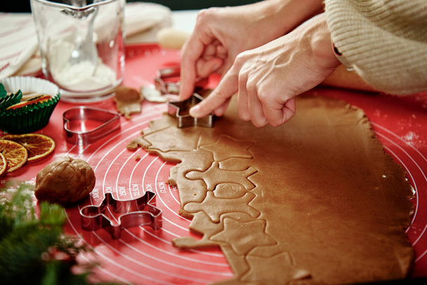 Γυναίκα προετοιμασία μπισκότα μελόψωμο στην κουζίνα με χριστουγεννιάτικα στολίδια. Γυναικεία χέρια που κόβουν τη ζύμη τζίντζερ με κόφτη για χειμερινές διακοπές - Φωτογραφία, εικόνα