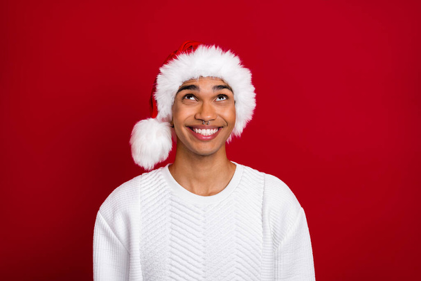Photo of young santa claus guy wear funny hat toothy beaming smile look up interested promo winter season sale isolated on red color background. - Photo, image