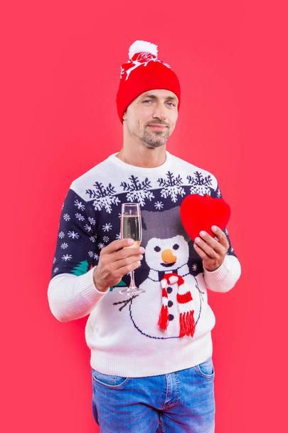 Nowy rok człowiek uśmiech odizolowany na czerwonym tle. Człowiek trzymać szampana na Nowy Rok w studio. Prezent na Nowy Rok i mężczyzna w swetrze. mężczyzna w kapeluszu noworocznym z szampanem i sercem. - Zdjęcie, obraz