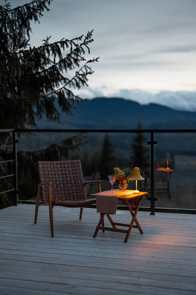 Spacious terrace with cozy table, chair and lamp highly in mountains at dusk. Cozy lounge place for rest at house on nature - Photo, Image