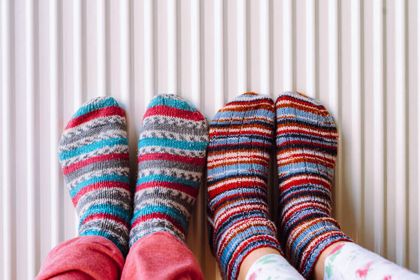 Two pairs of legs in knitted socks made of multi-colored woolen threads on heating radiator. Warm your feet near autonomous heater at home - Zdjęcie, obraz