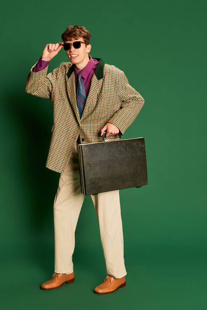 Studioaufnahmen junger Mann im Kostüm im Vintage-Stil, Anzug, der isoliert auf dunkelgrünem Hintergrund posiert. Konzept für Retro-Stil, Kreativität, Emotionen, Mimik, Mode und Business - Foto, Bild