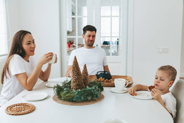 Family sitting at the table on a Christmas morning. Mother, father and their little son drinking tea and eating a cake for the Christmas breakfast. - 写真・画像