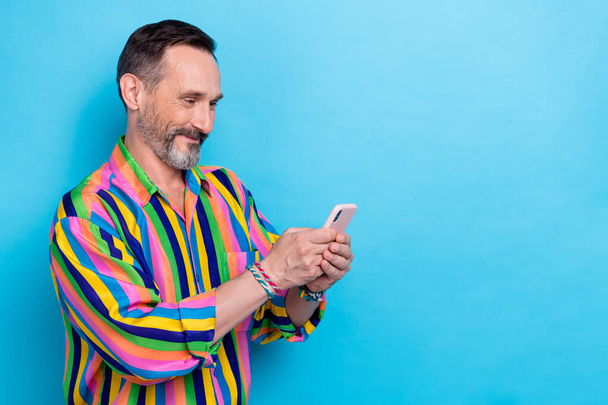 Photo of satisfied brunet hair mature age man wear striped colorful shirt hold his apple iphone choose eshop gift isolated on blue color background. - Foto, imagen