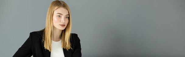 Blonde woman in black jacket looking away while standing near grey wall, banner  - Foto, afbeelding