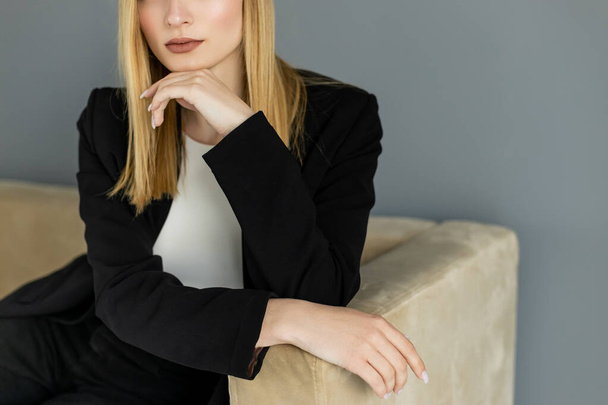 Cropped view of blonde woman in black jacket sitting on couch near grey wall  - Foto, Imagem