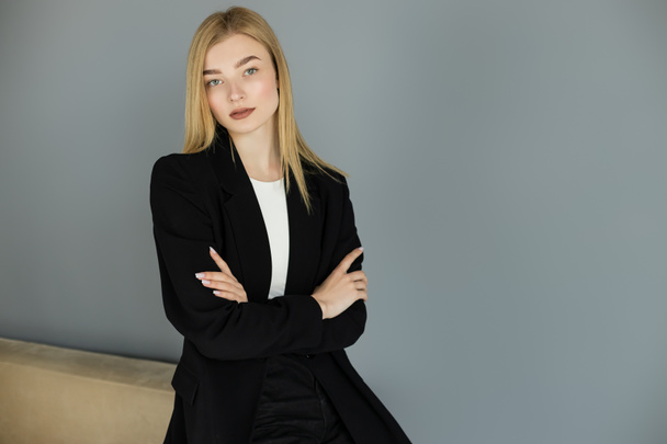 Confident woman in black jacket crossing arms near couch at home  - Photo, Image