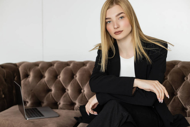 Portrait of blonde freelancer in jacket looking at camera near blurred laptop on velvet sofa  - 写真・画像