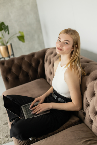 Pleased woman in top using laptop with blank screen on couch at home  - Foto, afbeelding