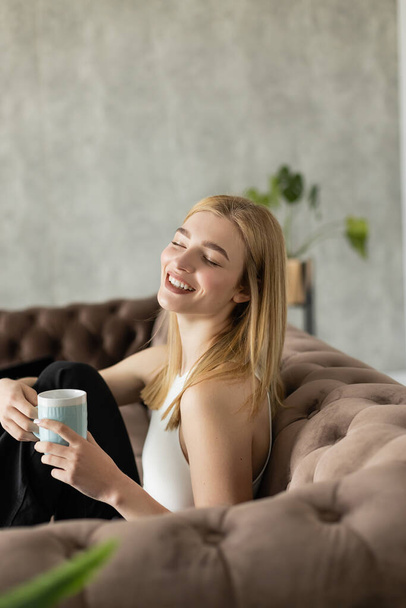 Junge blonde Frau lacht und hält Becher in der Hand, während sie zu Hause auf der Couch sitzt  - Foto, Bild