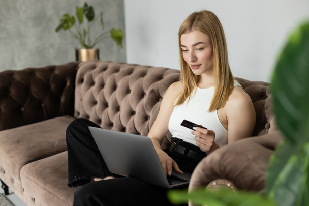 Mujer rubia positiva sosteniendo la tarjeta de crédito y el uso de ordenador portátil en el sofá moderno cerca de las plantas en casa  - Foto, imagen