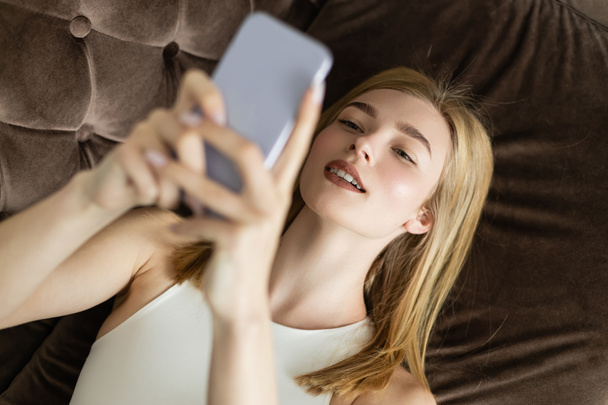 Vista dall'alto di una donna bionda sorridente che utilizza uno smartphone sfocato sul divano  - Foto, immagini