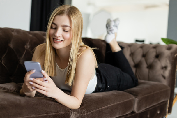 Задоволена блондинка використовує мобільний телефон, розслабляючись на дивані вдома
  - Фото, зображення
