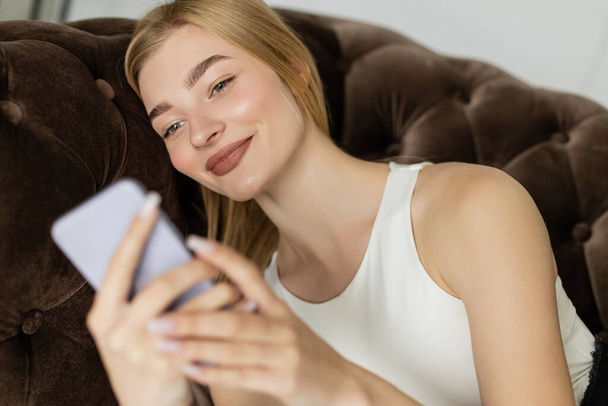 Mujer rubia feliz en la parte superior usando un teléfono inteligente borroso mientras se relaja en el sofá  - Foto, Imagen