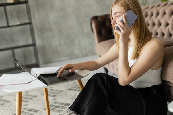 Блондинка тренер говорить на смартфоне и с помощью ноутбука в гостиной  - Фото, изображение