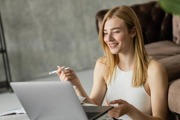 Ξανθιά γυναίκα χαμογελά κρατώντας στυλό κοντά σε φορητό υπολογιστή και σημειωματάρια κατά τη διάρκεια της online εκπαίδευσης  - Φωτογραφία, εικόνα