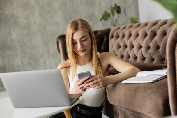 Hübsche Blondine nutzt Smartphone in der Nähe von Laptop und Notebook auf der Couch  - Foto, Bild