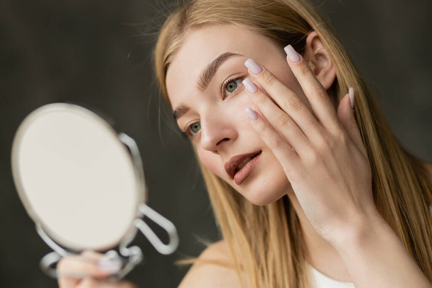 Молодая блондинка трогает лицо и держит размытое зеркало дома  - Фото, изображение