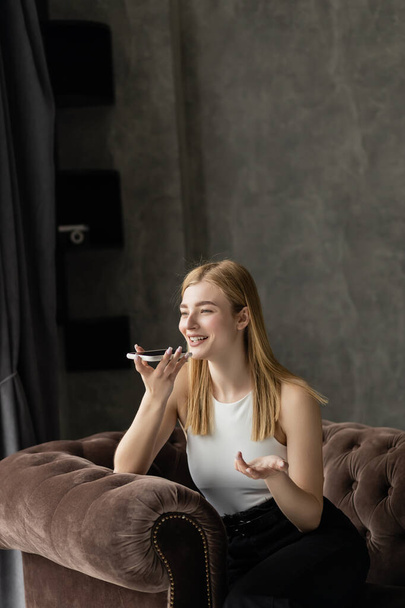 Smiling blonde woman recording voice message while sitting on armchair  - Fotó, kép