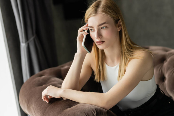 Evde koltukta otururken akıllı telefondan konuşan odaklanmış genç bir kadın.  - Fotoğraf, Görsel