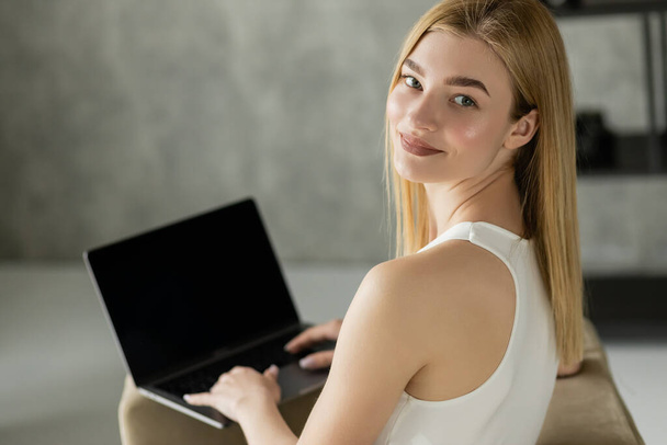 Θετική ξανθιά freelancer κοιτάζοντας κάμερα κοντά στο φορητό υπολογιστή με λευκή οθόνη στο σπίτι  - Φωτογραφία, εικόνα