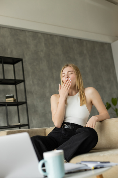 Блондинка фрилансер зевает возле размытых гаджетов и кофе в гостиной  - Фото, изображение