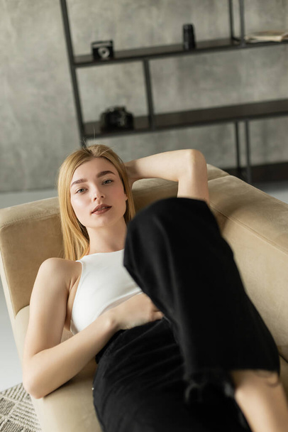 Kaunis nuori nainen päällä ja farkut katselee kameraa, kun makaa sohvalla kotona  - Valokuva, kuva