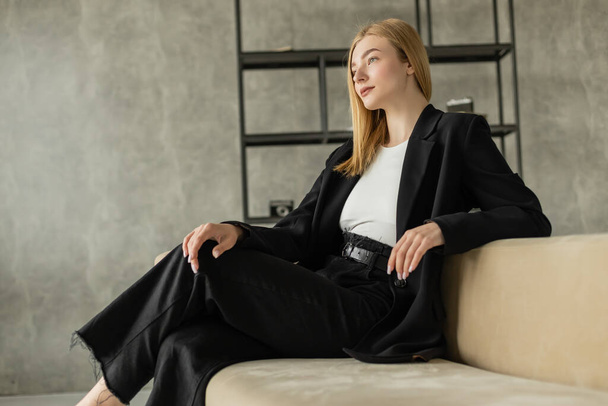 pensive blonde woman in black clothes sitting on couch in loft style apartment and looking away - Photo, Image