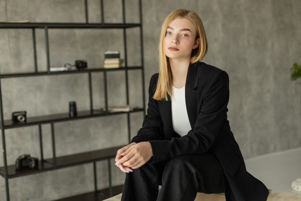 junge blonde Frau in schwarzem Blazer und schwarzer Hose sitzt in moderner Wohnung und blickt in die Kamera - Foto, Bild
