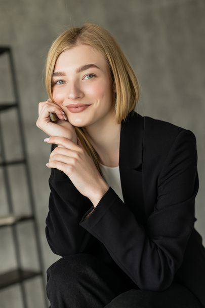 portrait of happy blonde woman in black blazer holding hand near face and smiling at camera at home - Photo, Image