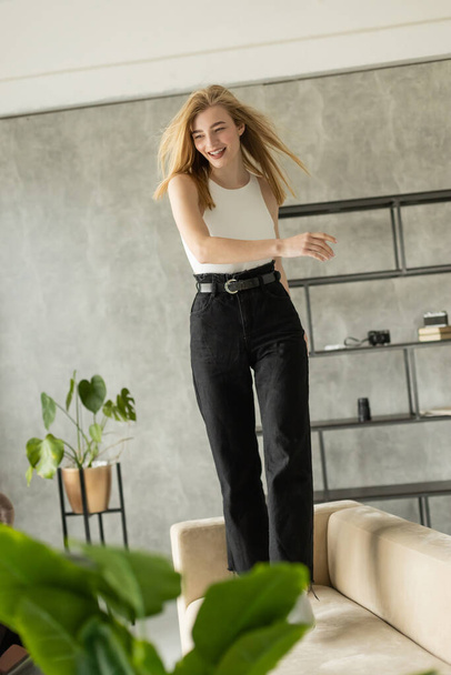 giovane donna eccitata in jeans neri che balla sul divano in appartamento moderno - Foto, immagini
