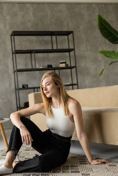 dreamy woman smiling and looking away while sitting on floor near couch in loft style living room - Fotografie, Obrázek