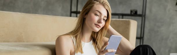 молода блондинка з мобільним телефоном, що сидить біля сучасного дивана вдома, банер
 - Фото, зображення
