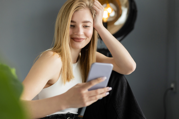 heureux femme blonde en débardeur blanc regardant flou téléphone mobile à la maison - Photo, image