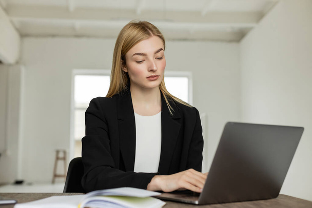 jovem treinador loira em blazer preto trabalhando no laptop perto de notebook borrado em casa - Foto, Imagem