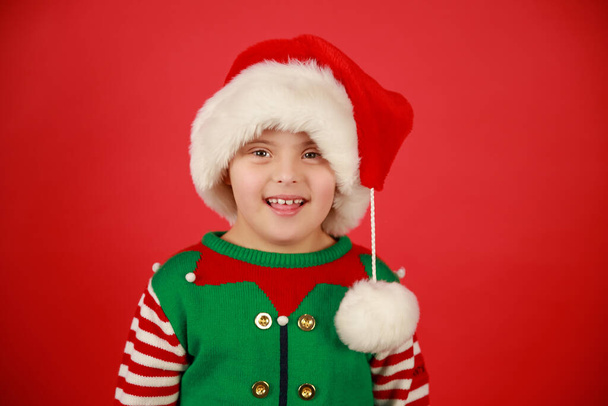 Niño feliz con síndrome de Down en un sombrero de Santa Claus y un suéter verde sobre un fondo rojo, humor navideño. Feliz Año Nuevo. - Foto, Imagen