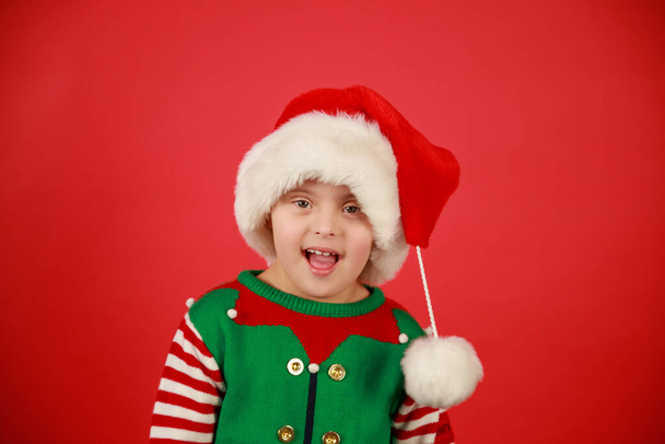 Happy dítě s Downovým syndromem v klobouku Santa Claus a zelený svetr na červeném pozadí, vánoční nálada. Šťastný nový rok. - Fotografie, Obrázek