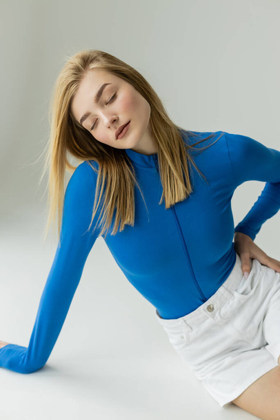 blonde woman in blue zipped turtleneck sitting with closed eyes and hand on hip on grey background  - Φωτογραφία, εικόνα