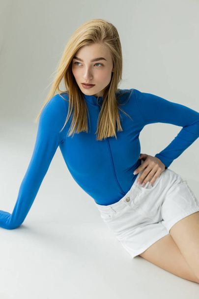 blonde woman in blue long sleeve shirt and white shorts looking away while posing with hand on hip on grey background  - Φωτογραφία, εικόνα
