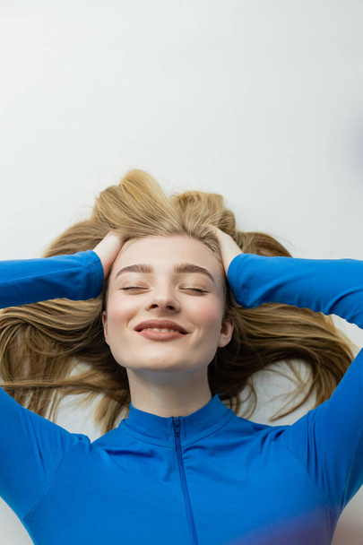 top view of young blonde woman smiling with closed eyes while lying on grey background  - Foto, Imagen