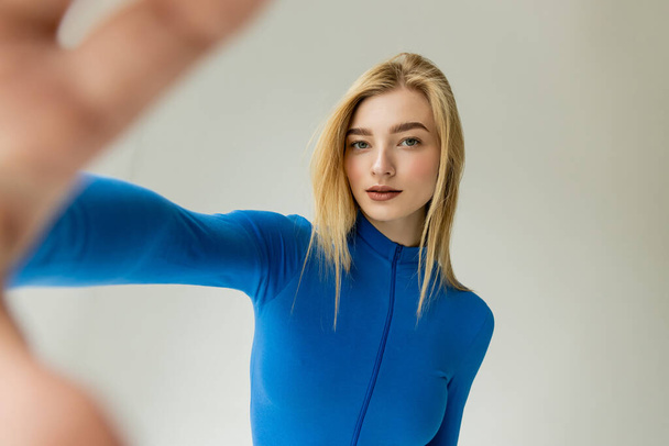 stylish blonde woman in blue turtleneck standing with blurred outstretched hand and looking at camera isolated on grey - Photo, Image
