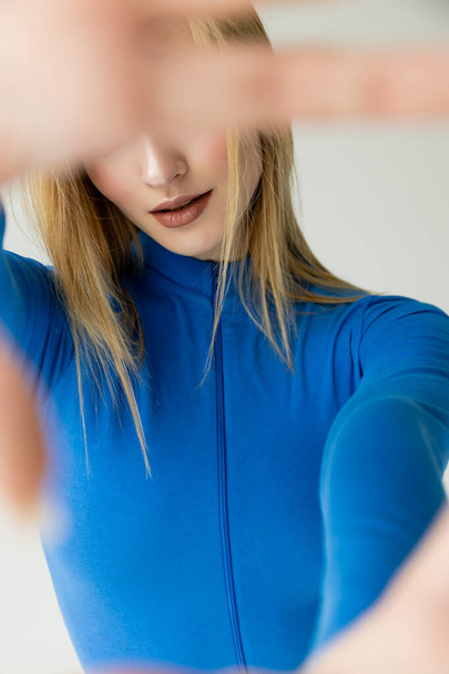 blonde woman in blue zipped turtleneck obscuring face with blurred hand isolated on grey - Photo, Image