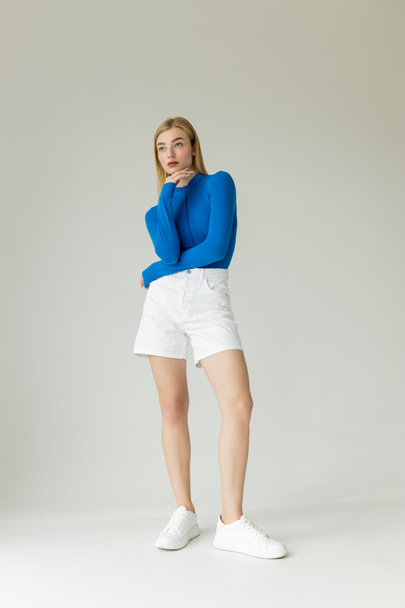 longitud completa de la esbelta mujer en camisa azul de manga larga y pantalones cortos blancos con zapatillas de deporte mirando hacia otro lado sobre fondo gris  - Foto, Imagen