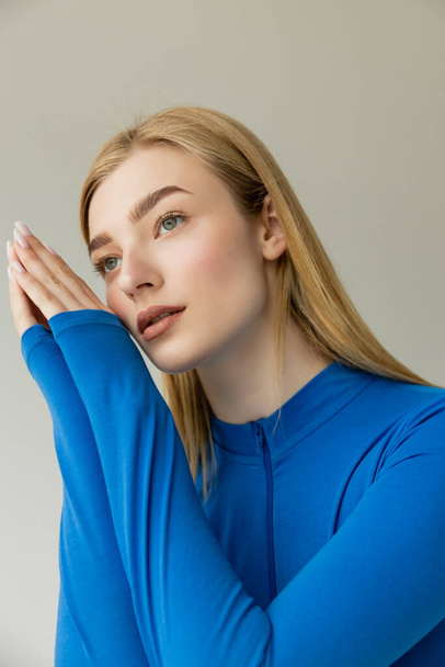 dreamy woman in blue long sleeve shirt holding hands near face and looking away isolated on grey - Photo, Image
