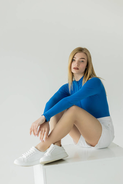 όμορφη γυναίκα σε μπλε μακρυμάνικο πουκάμισο και sneakers κάθεται σε λευκό κύβο και κοιτάζοντας κάμερα απομονώνονται σε γκρι - Φωτογραφία, εικόνα