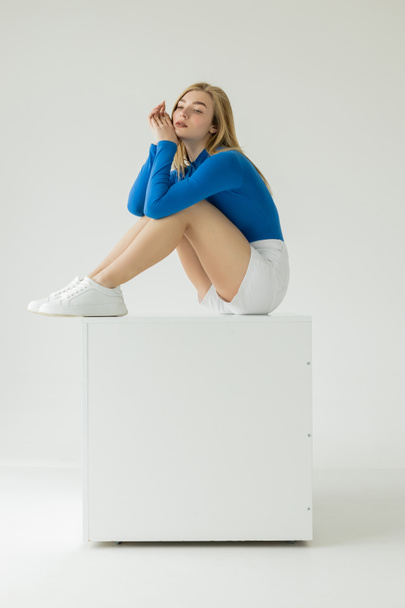 thoughtful woman in white shorts and blue turtleneck sitting on white cube on grey background - Foto, Imagen