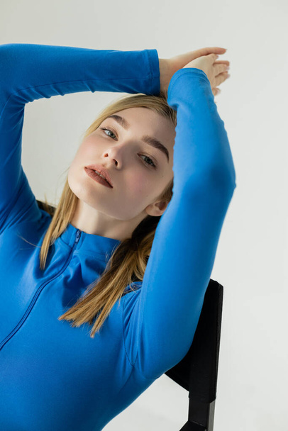junge Frau mit natürlichem Make-up trägt blauen Rollkragen, während sie mit den Händen über dem Kopf isoliert auf grau sitzt - Foto, Bild