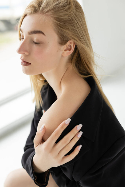 blonde woman in black blazer touching bare shoulder while posing with closed eyes at home - Foto, immagini