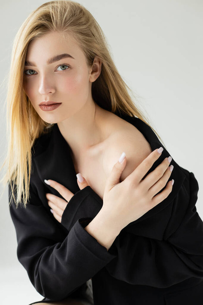sensual woman with blonde hair and naked shoulder posing in black jacket and looking at camera isolated on grey - Photo, image